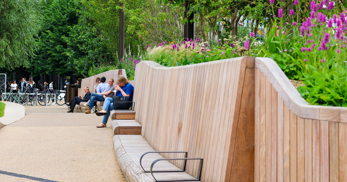 city scene with a collection of bikes and people standing with them to the left hand side, right hand side of image features a long run of wooden seating against a raised planter containing lots of flowers and plants