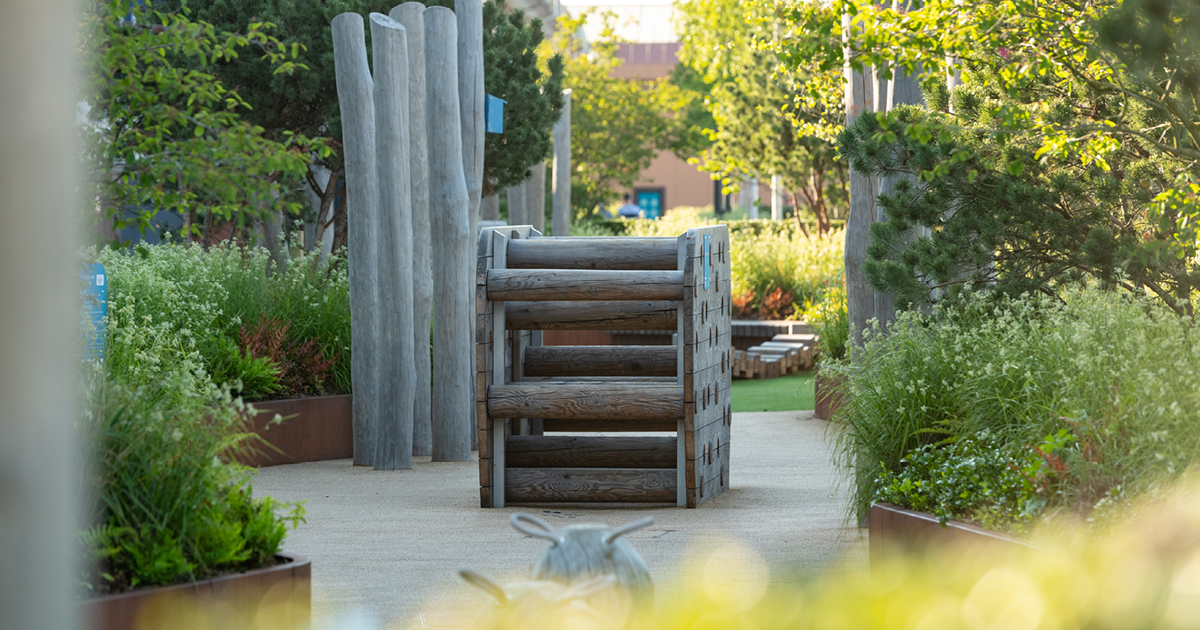 Outside shopping centre space between car park and store fronts. Wooden childrens climbing structure, plants and shrubbery around edge of image.