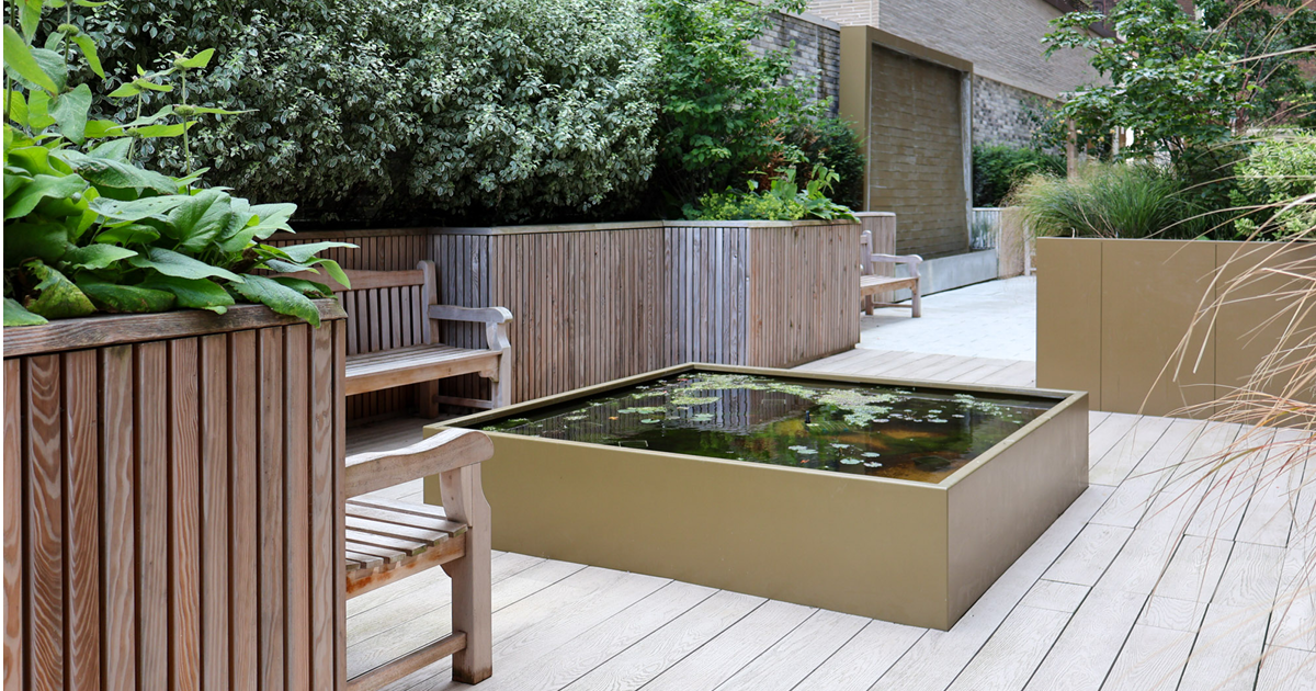 courtyard garden with a square pond in centre and timber benches around. Raised planter beds filled with foliage.