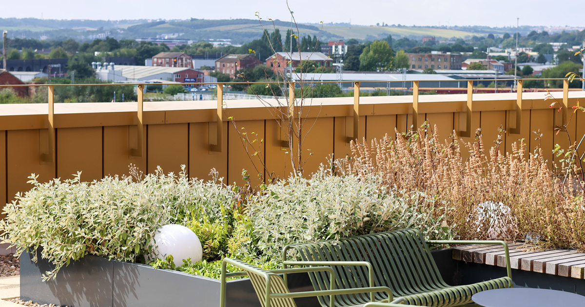 Roof top terrace with a view of city in background. Metal balustrade with industrial style. In foreground there is a green metal bench and chair surrounded by plants in grey PPC raised planters 