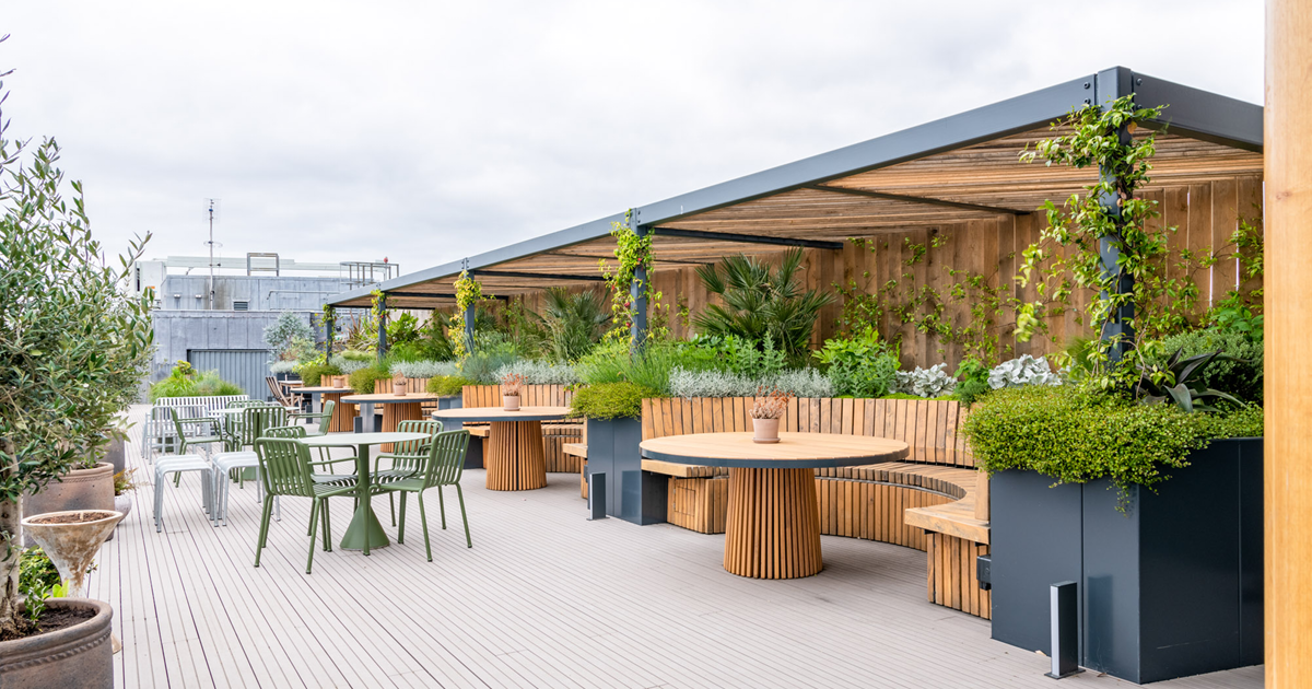 Roof top terrace with view of industrial building in the background. To the right of image there is a long pergola construction with metal framework and wood with a roof. Built into this there are 4 curved, wooded seating areas with matching round wooden tables. The floor is light green wooden deck and there are additional green and grey metal tables with chairs and benches. Green planting surrounds the space in raised planters and separate potted plants.