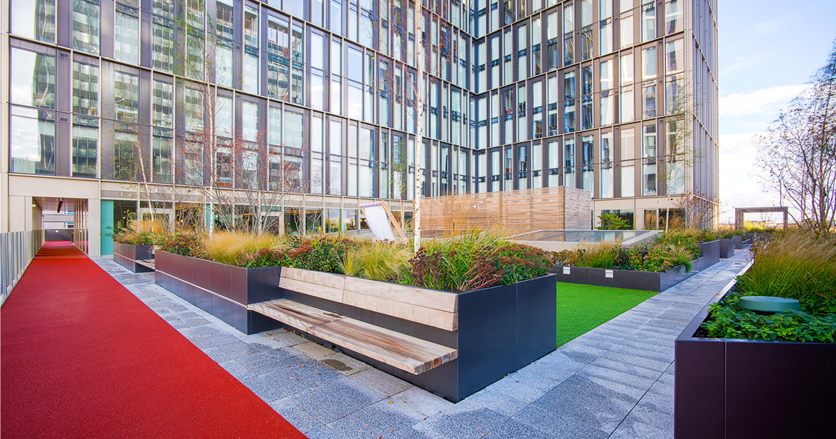 Roof top garden with multi level building in background. Foreground features a number of raised planters filled with plants and integrated seating. There is a red running track that loops around the garden.